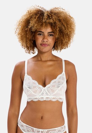 Femme aux cheveux bouclés portant un soutien-gorge en dentelle blanche et une lingerie assortie, debout devant un fond clair uni.