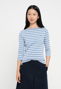 Ριγέ μπλούζα με μακριά μανίκια σε ανοιχτό μπλε και λευκό, με μποτ ντεκολτέ και μικρό λογότυπο, σε συνδυασμό με μια ναυτική φούστα με χρυσά κουμπιά.