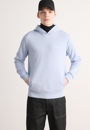 Lyseblå hettegenser med en jevn tekstur og logo på brystet, kombinert med mørk denimbukser med patchwork-detaljer.