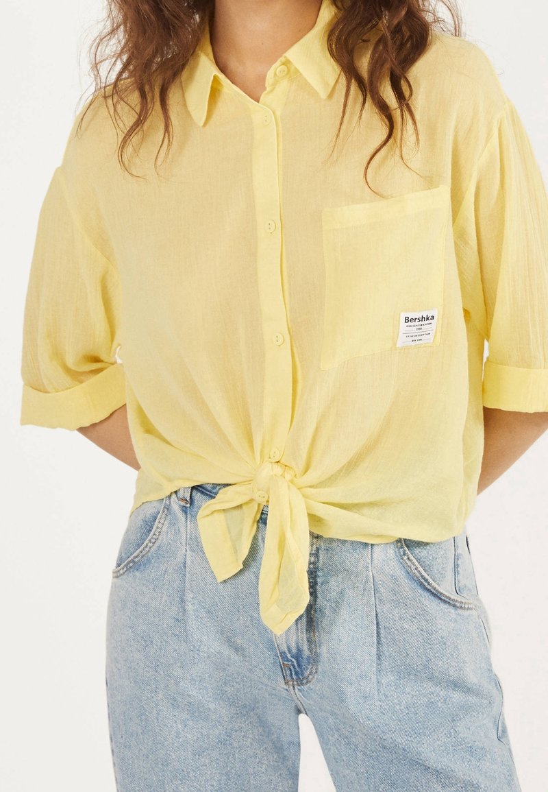 Femme portant une chemise jaune clair boutonnée, nouée à la taille avec une poche avant et un jean taille haute bleu clair.