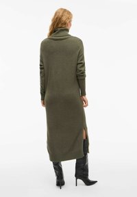 Robe en tricot vert olive avec col roulé, manches longues, fentes latérales et détails côtelés aux poignets, associée à des bottes noires à talons hautes arrivant jusqu'aux genoux.