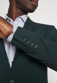Sötétzöld blézer sima textúrával, egygombos kialakítással, négy fekete gombbal az ujjakon, világoskék ing felett viselve.