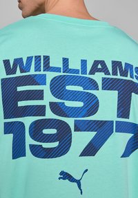 Jasnoniebieska bawełniana koszulka z ciemnoniebieskim napisem "WILLIAMS EST 1977" w pogrubionej, paskowanej czcionce, z małym logo Pumy w prawym dolnym rogu.