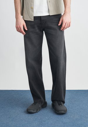 Blugi negri cu picior drept, cu o textură ușor decolorată, asortați cu pantofi negri fără șireturi. Fundalul este o mochetă albastră.