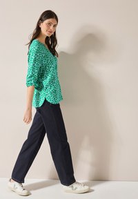 Grön blus med blommönster och trekvartsärmar, i kombination med mörka jeans och vita sneakers med gröna detaljer.