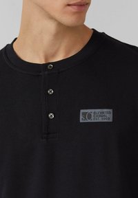 Zwarte geribde henley-shirt met een ronde kraag en drie zwarte knopen. Bevat een grijs label met "s.Oliver Elevated Casual Est. 1969."
