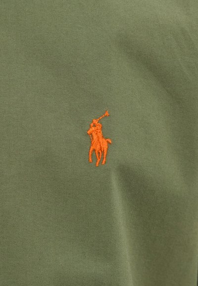 Alyvuogių žalios spalvos audinys su subtilia tekstūra, kairėje pusėje esantis oranžinės spalvos siuvinėtas arklio logotipas. Kasdieniško polo marškinėlių dizainas.