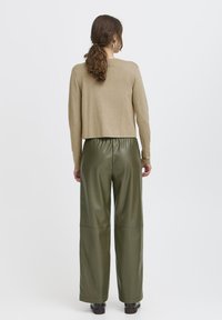 Beige, bokáig érő pulóver, hosszú ujjakkal, olajzöld, széles lábú műbőr nadrággal párosítva, sötét lábbelivel kiegészítve.