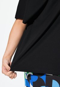 Černý krátký rukáv top s hladkým materiálem; ruka drží lem. Barevné vzorované látky sukně jsou viditelné níže.