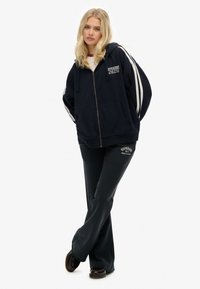 Sudadera con cremallera de color marino con rayas blancas en las mangas, que cuenta con un bolsillo tipo canguro y el logo "Superdry Athletic". Combinada con pantalones de chándal oscuros a juego.