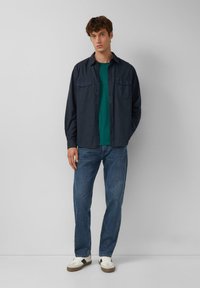 Camicia a maniche lunghe blu navy con due tasche sul petto sopra una t-shirt verde, abbinata a jeans blu e sneakers bianche con dettagli grigi.