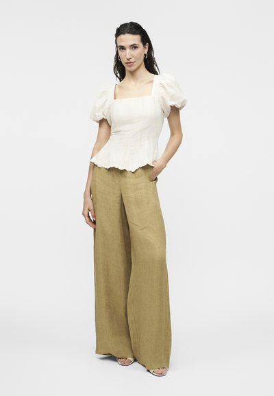 Γυναίκα με λευκό τοπ με τετράγωνη λαιμόκοψη και μανίκια-φουσκωτά, και μπεζ παντελόνι με φαρδιά πόδια, στέκεται με το ένα χέρι στην τσέπη της.