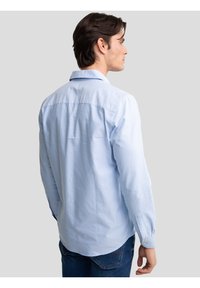 Mann mit dunklen Haaren, der ein hellblaues Langarmhemd und dunkle Jeans trägt, von hinten vor einem schlichten hellgrauen Hintergrund.