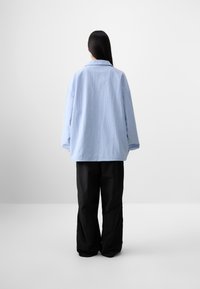 A person stands with their back to the camera, wearing a light blue oversized shirt and black pants, against a plain white background.