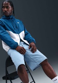 Homme aux cheveux tressés portant une veste Nike bleue et blanche et un short en denim clair, assis sur une chaise pliante noire devant un fond sombre.