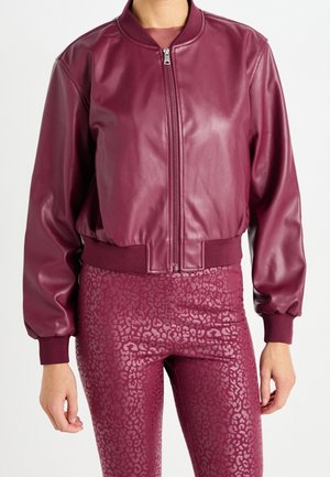 Personne portant une veste bomber en cuir synthétique bordeaux avec fermeture éclair et des leggings taille haute assortis avec un motif léopard subtil.