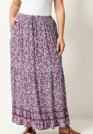 Kvinde iført en lang nederdel med blomstermønster i lilla og pink nuancer og en hvid top, hænderne hviler nær lommerne. Brune sandaler synlige.