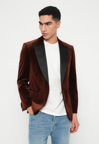 Man in een bruin fluwelen blazer met zwarte revers, een wit t-shirt en lichtblauwe jeans, kijkt naar opzij met één hand in zijn zak.