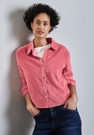 Mujer con cabello rizado se apoya en una pared gris, vestida con una camisa de botones de pana rosa sobre una camiseta blanca y jeans azul oscuro, con las manos en los bolsillos.