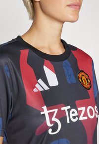 Čierny športový dres adidas s viacfarebným abstraktným vzorom v červenej a modrej. Obsahuje erb Manchester United a biely logotyp Tezos.