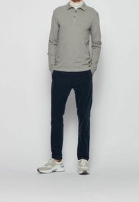 Man som bär en grå långärmad polo, vit polotröja, marinblå kordbyxor och vita sneakers, stående med händerna i fickorna.