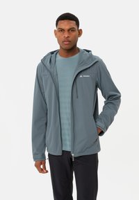 Veste softshell en teal avec fermeture éclair sur le devant, capuche ajustable et logo réfléchissant. Associée à une chemise à motifs bleu clair.