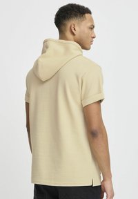 Beige kortærmet hættebluse med et struktureret, vaffelmønstret design. Har opbundne ærmer og sideslids i bunden.