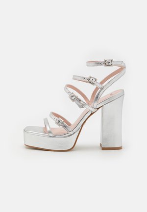 Hõbedased platvormsandaalid, millel on mitmekesised õhukesed rihmad ja ruudukujuline konts. Omab läikivat metalse viimistlust ja pandlad rihmade kaunistamiseks.