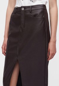 Femme portant une jupe en similicuir marron foncé taille haute avec une fente à l'avant et un top blanc rentré.