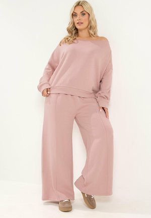 Blond kvinde står iført en løs, lyserød off-shoulder sweatshirt og vidde bukser med beige sneakers mod hvid baggrund.