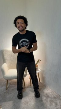 Άνδρας που χαμογελά με σταυρωμένα χέρια, φοράει μαύρη μπλούζα Rip Curl Hawaii και σκουρόχρωμο παντελόνι, στέκεται κοντά σε λευκή καρέκλα και μικρό τραπέζι με φυτό.