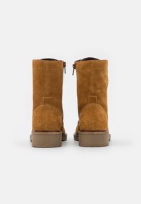 Botas de tornozelo em camurça castanha clara com um fecho lateral, detalhe no salto curvo e solas de borracha planas. Textura suave e design minimalista.