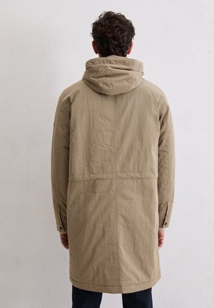 Uomo con capelli corti ricci che indossa una lunga giacca beige con cappuccio, in piedi di fronte a un muro bianco uniforme, con le mani lungo i fianchi.