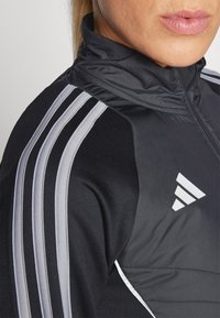Zwarte sportjack met hoge kraag, voorzien van grijze accenten met drie strepen en een wit logo op de borst. Glad, lichtgewicht materiaal.