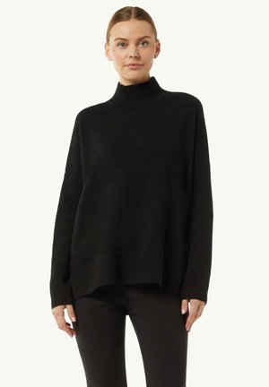 Schwarzer Rollkragenpullover mit gerippten Ärmeln und Saum, mit lockerer Passform und strukturiertem Stoff. Getragen mit schwarzen Hosen.