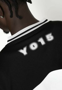 Schwarzes Trikot mit einem weißen Kragen und weißen Akzenten. Auf der Rückseite befindet sich die Aufschrift "YO15" in gepunktetem Druck. Weicher Stoff mit strukturiertem Finish.