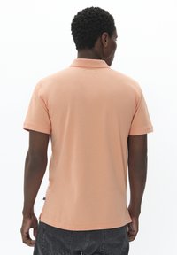 Polo tričko s krátkým rukávem v broskvovém bavlněném materiálu, s klasickým límcem, bočními rozparky a prošitými lemy. Jednoduchý design, bez vzorů nebo ozdob.