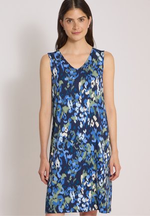 Femme portant une robe sans manches avec un motif floral abstrait bleu, vert et blanc sur un fond clair uni.