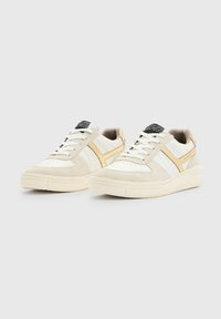 Białe i beżowe sneakersy o zamszowej fakturze, z perforacjami, kontrastującymi paskami i grubą gumową podeszwą. Akcentowane czarną metką.