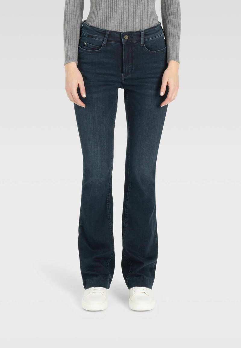 Femme portant un jean bootcut bleu foncé, un pull gris côtelé et des baskets blanches, debout sur un fond uni gris clair.