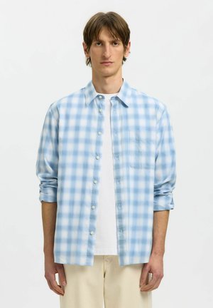 Junger Mann trägt ein hellblau-weiß kariertes Hemd über einem weißen T-Shirt, mit beigen Hosen, steht vor einem schlichten Hintergrund.