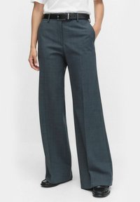 Graue weite Hosen aus strukturiertem Stoff mit einer mittelhohen Taille und einem schwarzen Ledergürtel, kombiniert mit schwarzem Schuhwerk.