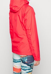 Veste à capuche rouge vif avec une texture légère et lisse. Portée avec des leggings colorés à motifs comprenant des dessins bleus, orange et gris.