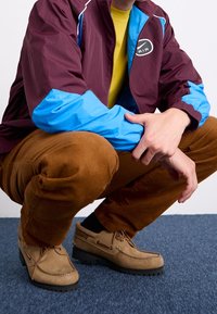 Tummanpunainen ja sininen vetoketjullinen takki, keltainen paita, ruskeat puuvillahousut ja beiget nahkakengät, joissa ovat uritetut pohjat ja solmitut nauhat.