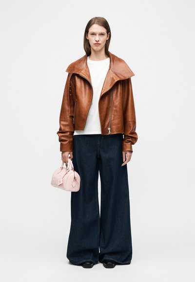 Γυναίκα που φοράει καφέ δερμάτινο μπουφάν, λευκό πουκάμισο, φαρδιά σκούρα μπλε τζιν, μαύρα παπούτσια, κρατώντας μικρό ανοιχτό ροζ τσαντάκι, στέκεται σε λευκό φόντο.