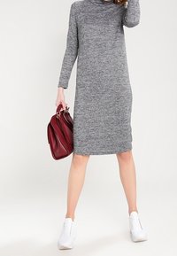 Graues Strickkleid mit hohem Kragen und langen Ärmeln, kombiniert mit weißen Turnschuhen und einer roten Handtasche. Glatte Textur. Oberschenkellang.