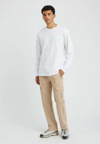 Chemise blanche à manches longues avec un logo sur la poitrine, associée à un pantalon cargo beige doté de plusieurs poches et des chaussures décontractées.