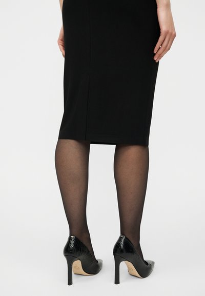 Γυναίκα εμφανίζεται από πίσω φορώντας μαύρες διαφανείς καλσόν, μαύρη φούστα μήκους έως το γόνατο και μαύρα ψηλοτάκουνα παπούτσια με υφασματικό μοτίβο.