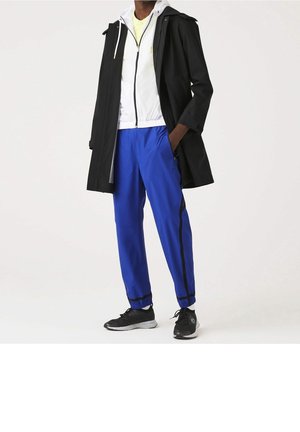 Musta vedenpitävä takki valkoisen vetoketjullisen jakun päällä, yhdistettynä sinisiin urheiluhousuihin, joissa on mustat sivuraitat ja mustat lenkkarit.