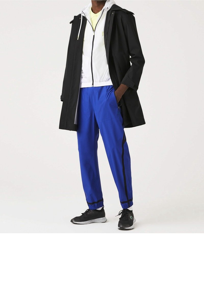 Černý voděodolný kabát přes bílou bundu na zip, doplněné modrými sportovními kalhotami s černými bočními pruhy a černými teniskami.